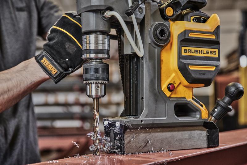 A person using a DEWALT brushless magnetic drill press to drill into a metal surface. Metal shavings are visible around the drill bit, and the person is wearing a black DEWALT glove.